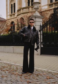 Czarna skórzana kurtka z futrzanym kołnierzem, szary sweter, wzorzyste krawat oraz czarne rozszerzane spodnie. Dodatki to okulary przeciwsłoneczne i rękawiczki.