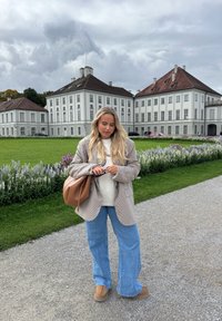 Ruskea ruutukuvioinen ylipitkä bleiseri, kermaan vivahtava neule, vaaleansiniset leveälahkeiset farkut, ruskea käsilaukku ja beigejä saappaat, seisomassa kukkien ympäröimässä puutarhassa.