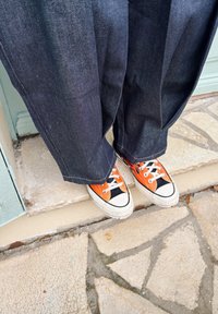 Personne debout sur un trottoir en pierre, portant un jean large foncé et des baskets en toile orange vif avec des lacets et semelles blanches.