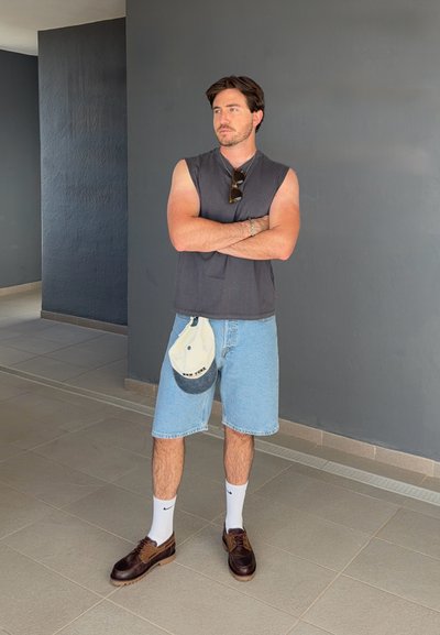 Hombre de pie con los brazos cruzados, vistiendo una camiseta negra sin mangas, pantalones cortos de mezclilla, calcetines blancos, zapatos marrones y sosteniendo una gorra.