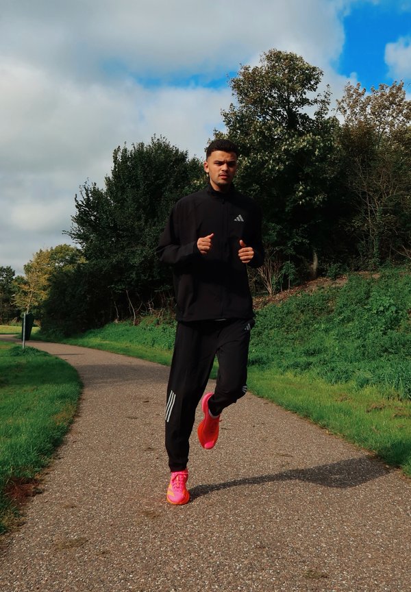 Un hombre vestido con ropa deportiva negra y zapatillas de color rosa intenso corre por un sendero pavimentado en un parque verde.