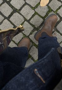 Brązowe skórzane botki z gumowymi podeszwami, noszone z ciemnoniebieskimi dżinsami. Obok na brukowanej ścieżce leży wzorzysty worek z widocznym złotym zamkiem.