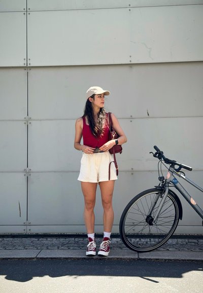 Una mujer con una blusa burdeos y pantalones cortos blancos está al lado de una bicicleta gris. Lleva una gorra beige y zapatillas rojas, sosteniendo un bolso pequeño.