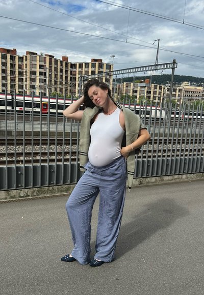 Mono sin mangas blanco, chal gris oversized, pantalones de pierna ancha a rayas azules y zapatos de tela azul, de pie cerca de las vías del tren con edificios de fondo.