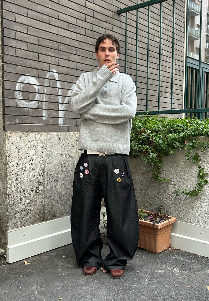 Gray knitted sweater with a collared neckline, paired with black wide-legged trousers featuring multiple colorful buttons. Brown shoes, urban background.