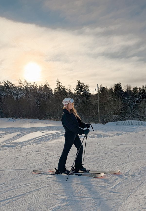 Vrouw in een zwarte skipak met helm en skibril staat op ski's in de sneeuw met skistokken, bos en ondergaande zon op de achtergrond.