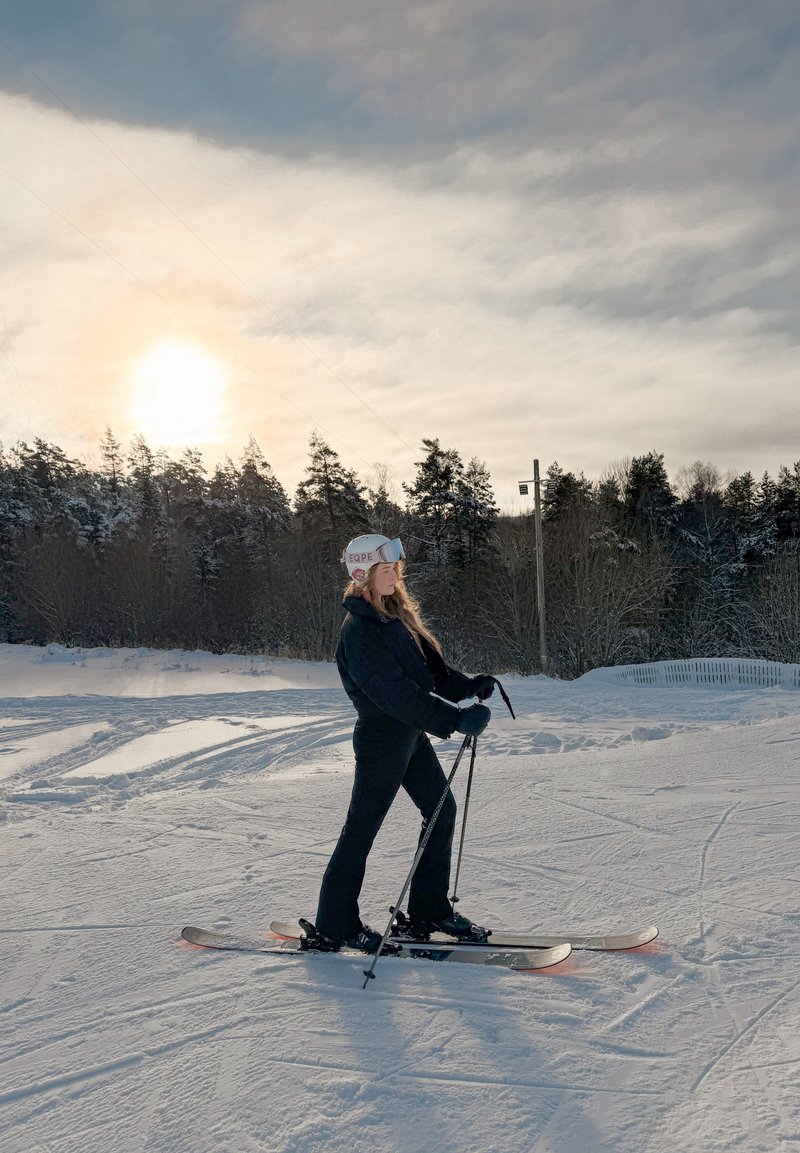 Femme en tenue de ski noire portant un casque et des lunettes de ski, debout sur des skis dans la neige avec des bâtons de ski, forêt et coucher de soleil en arrière-plan.