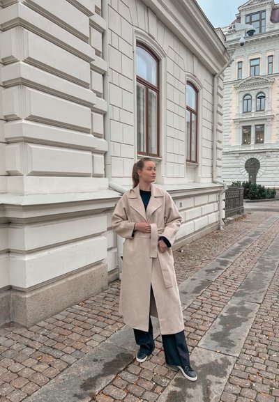 Abrigo largo beige con cinturón y amplias solapas, usado sobre una parte superior oscura. Combinado con pantalones marinos y zapatillas grises, en una calle adoquinada.