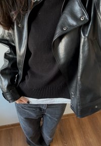 Veste en cuir noire avec matériel argenté, superposée sur un pull noir et une chemise blanche. Pantalon large gris foncé. Textures lisses.