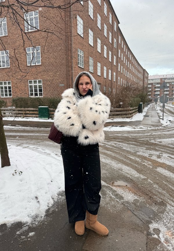Kvinna i en vit pälsjacka med fläckar och luva står på en snötäckt stadsgata nära röda tegelbyggnader under snöfall.