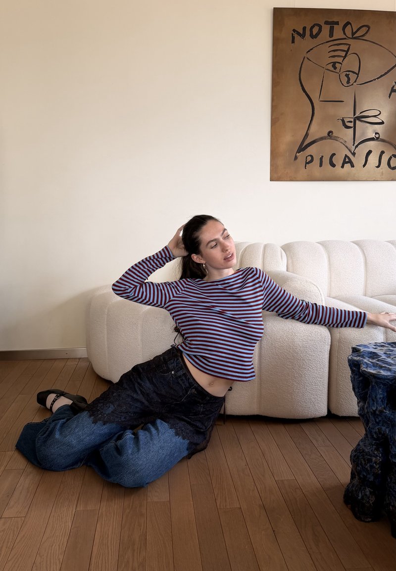 Femme en haut rayé et jean large allongée sur un sol en bois à côté d'un canapé blanc incurvé, tendant la main vers une table texturée bleu foncé.