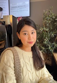 Pullover lavorato a maglia color avorio con motivo a treccia, manico di borsa intrecciato beige, ambientazione interna con piante e menù delle bevande sullo sfondo.