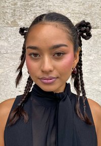 Jongedame met gevlochten haar in twee bollen gestyled, draagt een mouwloze zwarte top, met natuurlijke make-up en rode oorbellen, tegen een stenen muur.