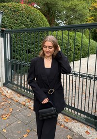 Černé sako na míru s širokým klopovým límcem a páskem v pase, doplněné o přiléhavý top a černou kabelku. Hladká textura kontrastuje s kamennou cestou.