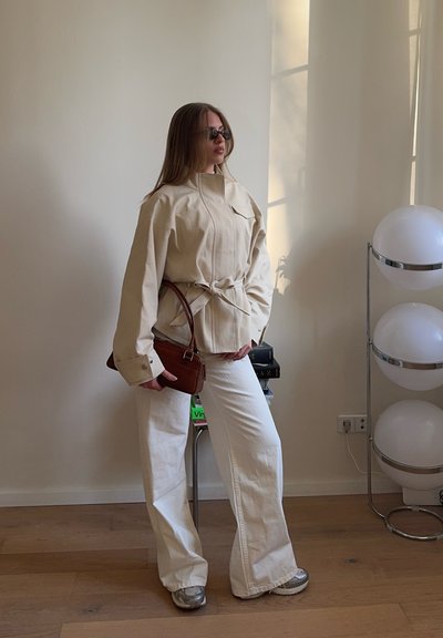 Mujer con chaqueta beige y pantalones blancos de pierna ancha, sosteniendo un bolso marrón, de pie junto a una lámpara blanca redonda en una habitación minimalista.