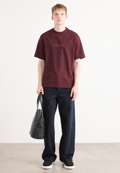 Camiseta de algodón burdeos con logo bordado, combinada con jeans de pierna ancha azul oscuro y zapatillas negras. El modelo sostiene una bolsa de tote negra.