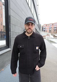 Άνδρας που φοράει μαύρο πλεκτό μπουφάν με φερμουάρ και μαύρο καπέλο, στέκεται σε χιονισμένο πεζοδρόμιο σε αστικό περιβάλλον κοντά σε σύγχρονα κτίρια.