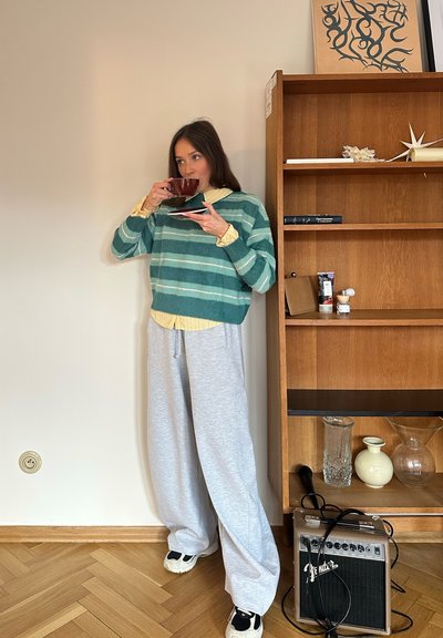 Suéter de felpa a rayas verdes sobre una camisa amarilla, pantalones de chándal grises, sosteniendo una taza de color rojo oscuro sobre un plato negro. Estantería de madera clara de fondo.