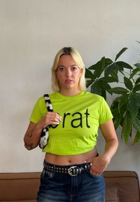 Svetla limonasto zelena skrajšana majica s črnim napisom "brat". Oblečena s hlačami iz jeansa z visokim pasom in črnim pasom s studenci. Črna in bela vrečka z vzorcem.