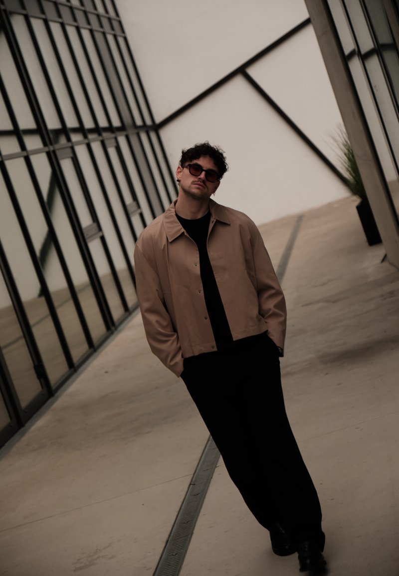 Giovane uomo con occhiali da sole, giacca beige e pantaloni neri che si inclina leggermente mentre cammina lungo un corridoio di un edificio moderno in vetro.