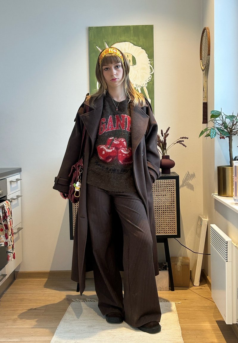 Jeune femme portant un long manteau marron et un pantalon rayé, un pull à motif cerise, un bandeau orange, debout dans une pièce avec des plantes et des œuvres d'art.