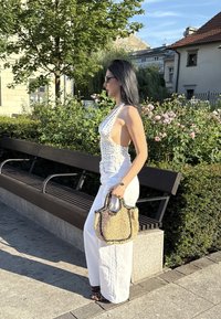Bijele široke hlače u kombinaciji s majicom s točkicama, upotpunjene pletenom torbom i cipelama s životinjskim uzorkom. Prirodni pozadinski elementi.