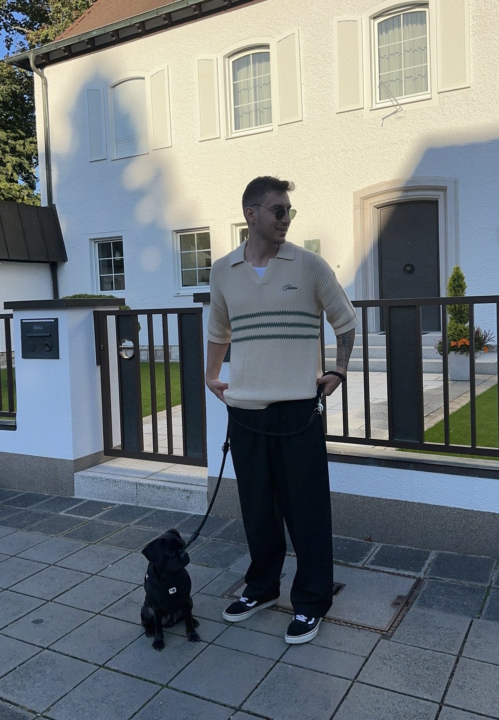 Bežs adīts polo ar īsām piedurknēm un zaļām svītrām, apvienots ar melniem platām biksēm un melniem sporta apaviem. Blakus sēž mazs melns suns pie pavadas.