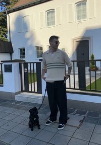 Bežs adīts polo ar īsām piedurknēm un zaļām svītrām, apvienots ar melniem platām biksēm un melniem sporta apaviem. Blakus sēž mazs melns suns pie pavadas.