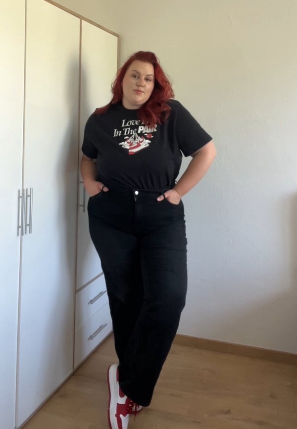 Jeans pretos de cintura alta com bolsos frontais, combinados com uma t-shirt preta com um design gráfico em vermelho e branco, e ténis vermelhos.