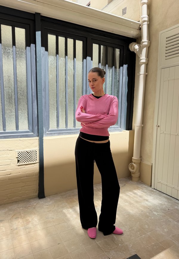 Jeune femme debout à l'intérieur, bras croisés, portant un pull rose, un pantalon noir et des chaussures roses, devant un mur vitré et des tuyaux beiges.