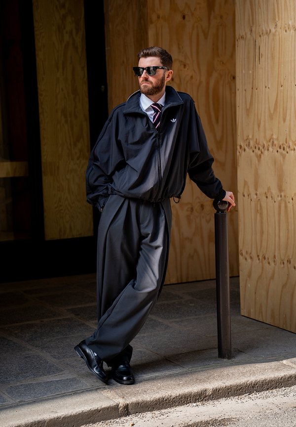 Man wearing sunglasses, a black jacket, and wide trousers leans against a black post on a city pavement in front of a wooden panel wall.