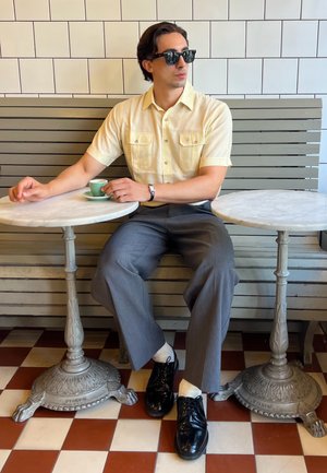 Camisa amarilla claro de manga corta con dos bolsillos en el pecho, pantalones grises y zapatos de vestir negros. Dos mesas con tapa de mármol y una taza de espresso verde.