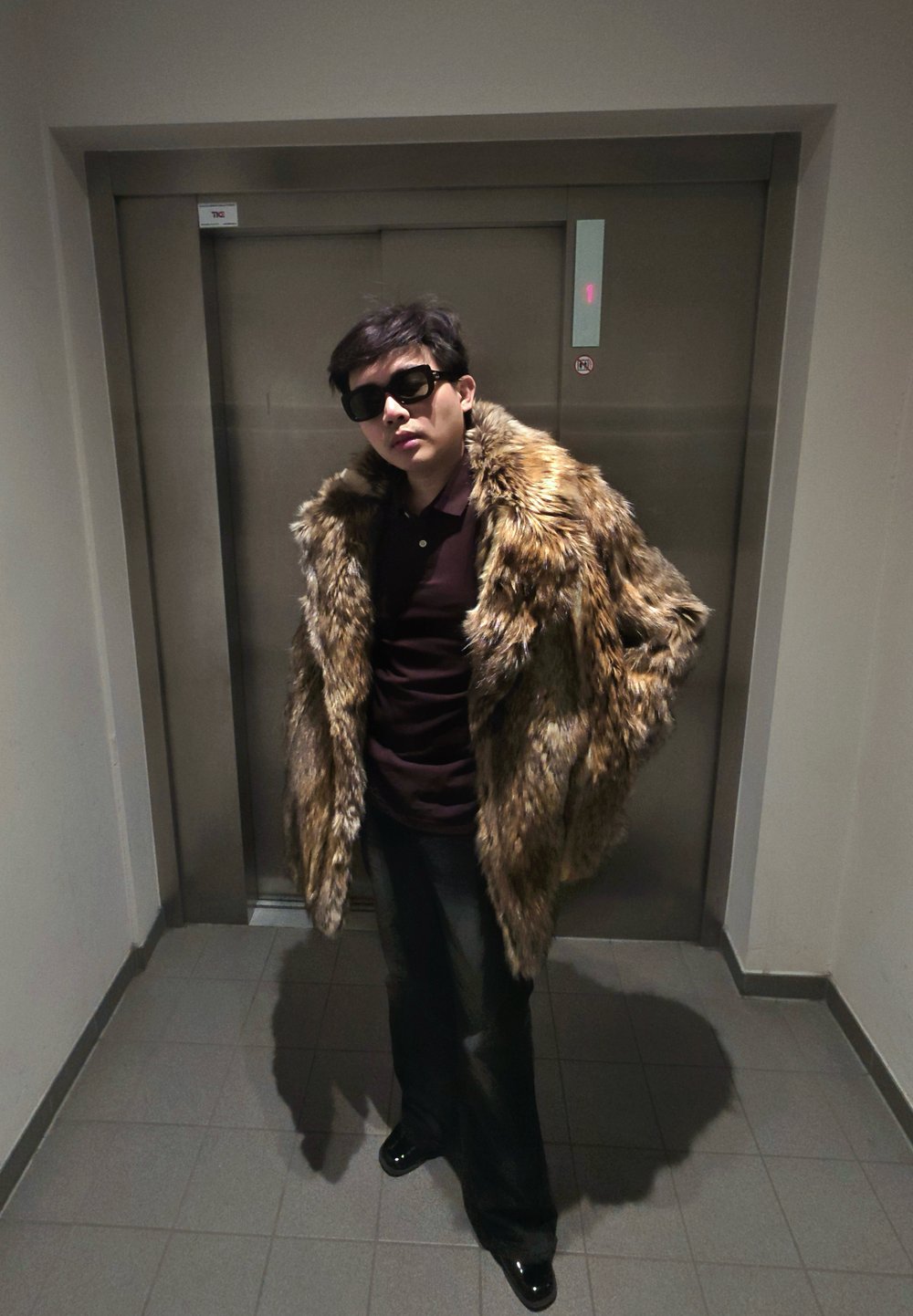 Homme portant des lunettes de soleil sombres, un pantalon noir, une chemise bordeaux et un grand manteau en fourrure marron se tient devant les portes fermées d'un ascenseur.