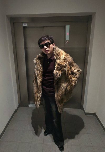 Hombre con gafas de sol oscuras, pantalones negros, camisa granate y un gran abrigo de piel marrón, de pie frente a las puertas cerradas de un ascensor.