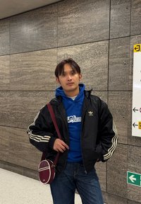 Veste noire avec des rayures blanches, sweat à capuche bleu, jean bleu et sac bandoulière bordeaux contre un mur gris texturé.