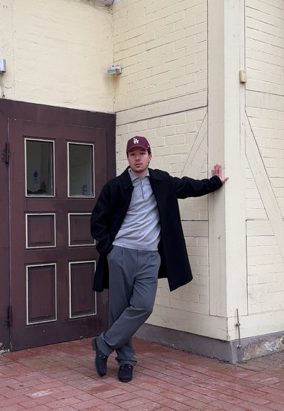 Joven con abrigo oscuro y gorra de béisbol apoyado contra una pared de ladrillos beige cerca de una puerta de madera marrón, de pie sobre un pavimento de ladrillos rojos.