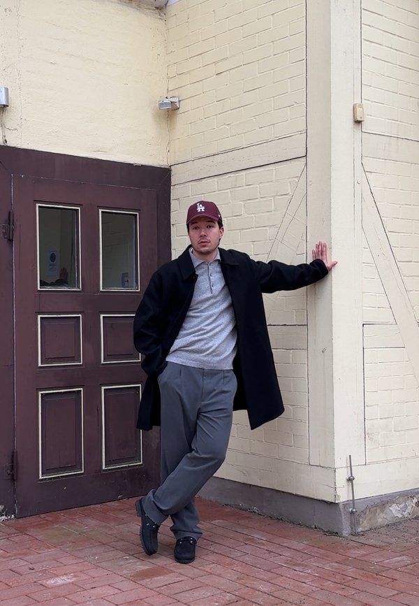 Mladý muž v tmavém kabátu a baseballové čepici opírající se o béžovou cihlovou zeď u hnědých dřevěných dveří, stojící na červené cihlové dlažbě.