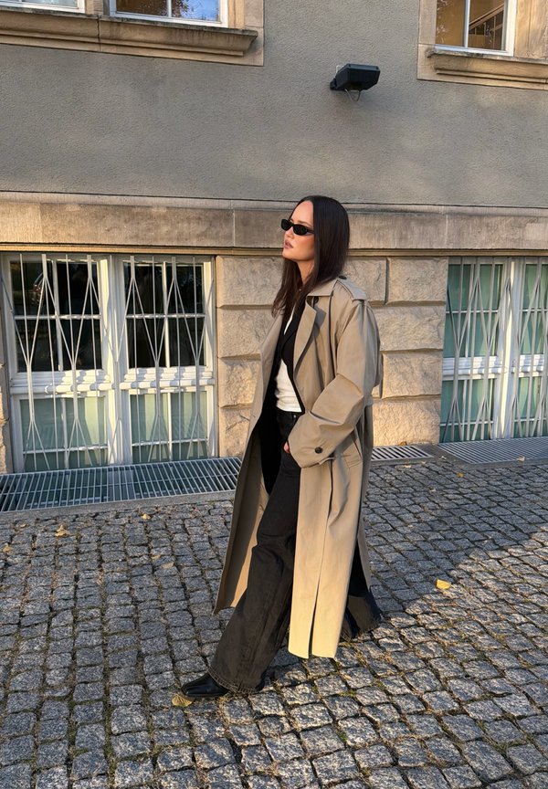 Una mujer con gafas de sol y una gabardina beige camina por una calle empedrada junto a un edificio de piedra con ventanas enrejadas.
