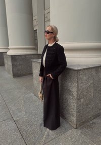 Schwarze kurze Jacke mit goldenen Knöpfen, weißem Oberteil und langem schwarzem Rock; ergänzt durch eine kleine gemusterte Handtasche und eine Sonnenbrille.