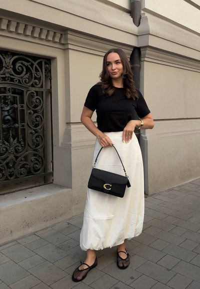 Top negro de manga corta, falda midi blanca y fluida, zapatos negros tipo slip-on de malla, y un bolso negro con herrajes dorados y detalles de logo.