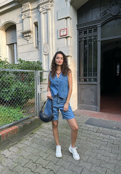 Chaleco de denim y shorts con botones, combinados con zapatillas blancas. Bolso negro con un diseño curvado. El fondo presenta vegetación y arquitectura.