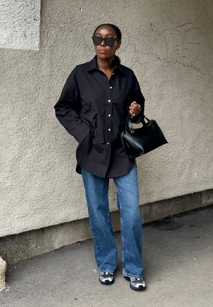 Mujer con camisa negra extragrande, vaqueros azules, zapatillas blancas y negras, gafas de sol negras y sosteniendo un bolso negro, de pie junto a una pared texturizada.