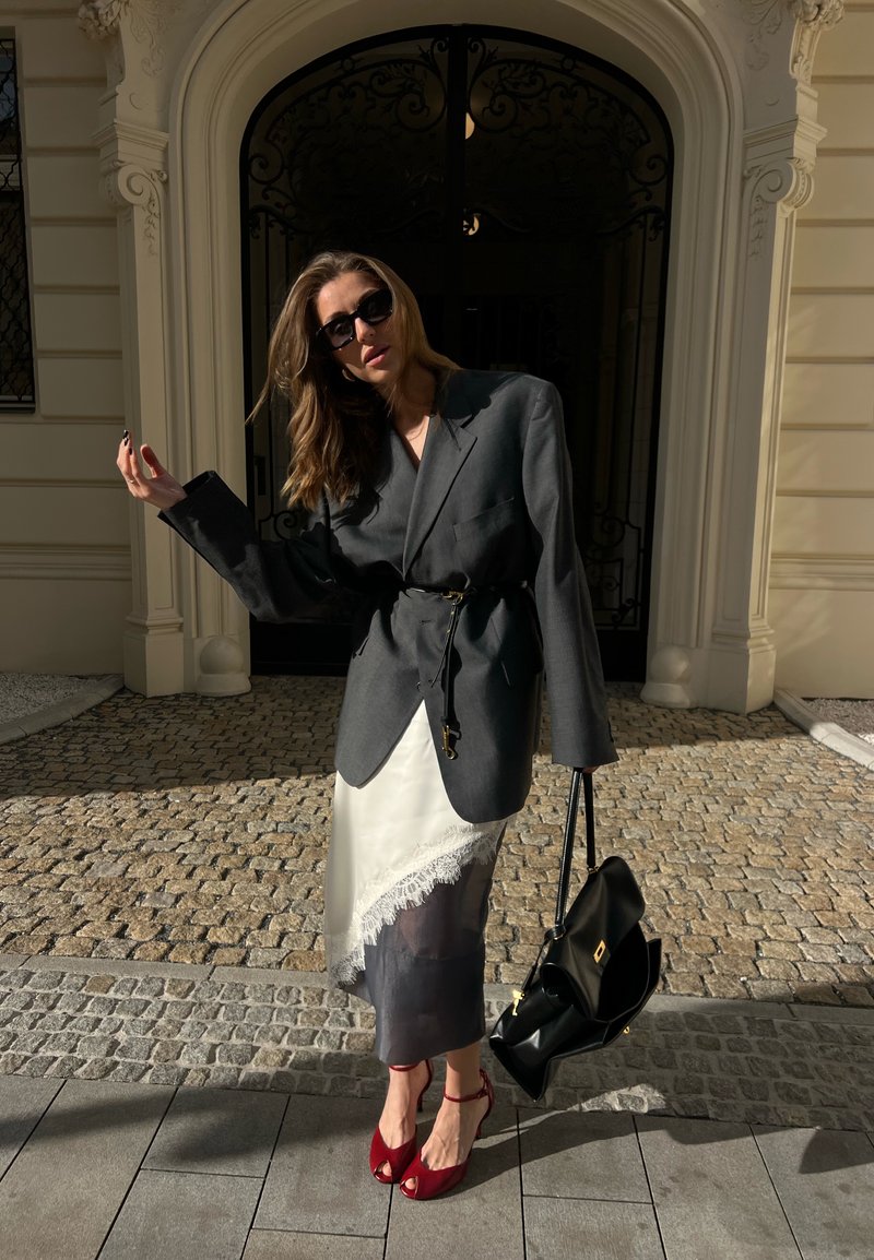 Femme en blazer foncé surdimensionné ceinturé à la taille, jupe en dentelle blanche superposée, talons rouges, lunettes de soleil, tenant un sac à main noir, debout sur une rue pavée.