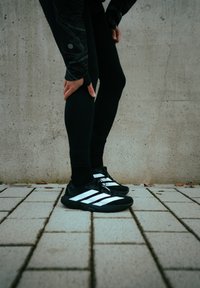 Chaussures de sport noires avec des bandes blanches, présentant une texture lisse et un design à lacets, portées avec un legging noir sur une surface pavée.