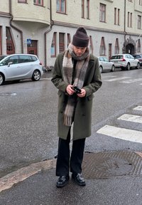 Homme portant un manteau olive, une écharpe à carreaux et un bonnet marron debout sur une rue mouillée près d'un passage pour piétons, regardant des gants dans ses mains.
