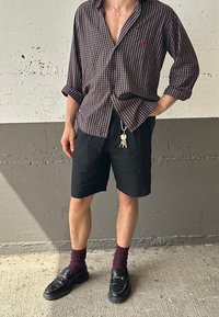 Camicia a quadri marrone e blu navy con maniche arrotolate, pantaloni corti neri, calzini bordeaux e mocassini neri con dettagli metallici. Chiavi attaccate.