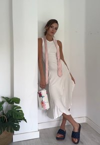 Weißes geripptes Kleid mit einer rosa Satin-Schleife, dazu eine mehrfarbige Blumenhandtasche. Marineblau gemusterte Sandalen. Indoor-Umgebung mit Pflanzen.