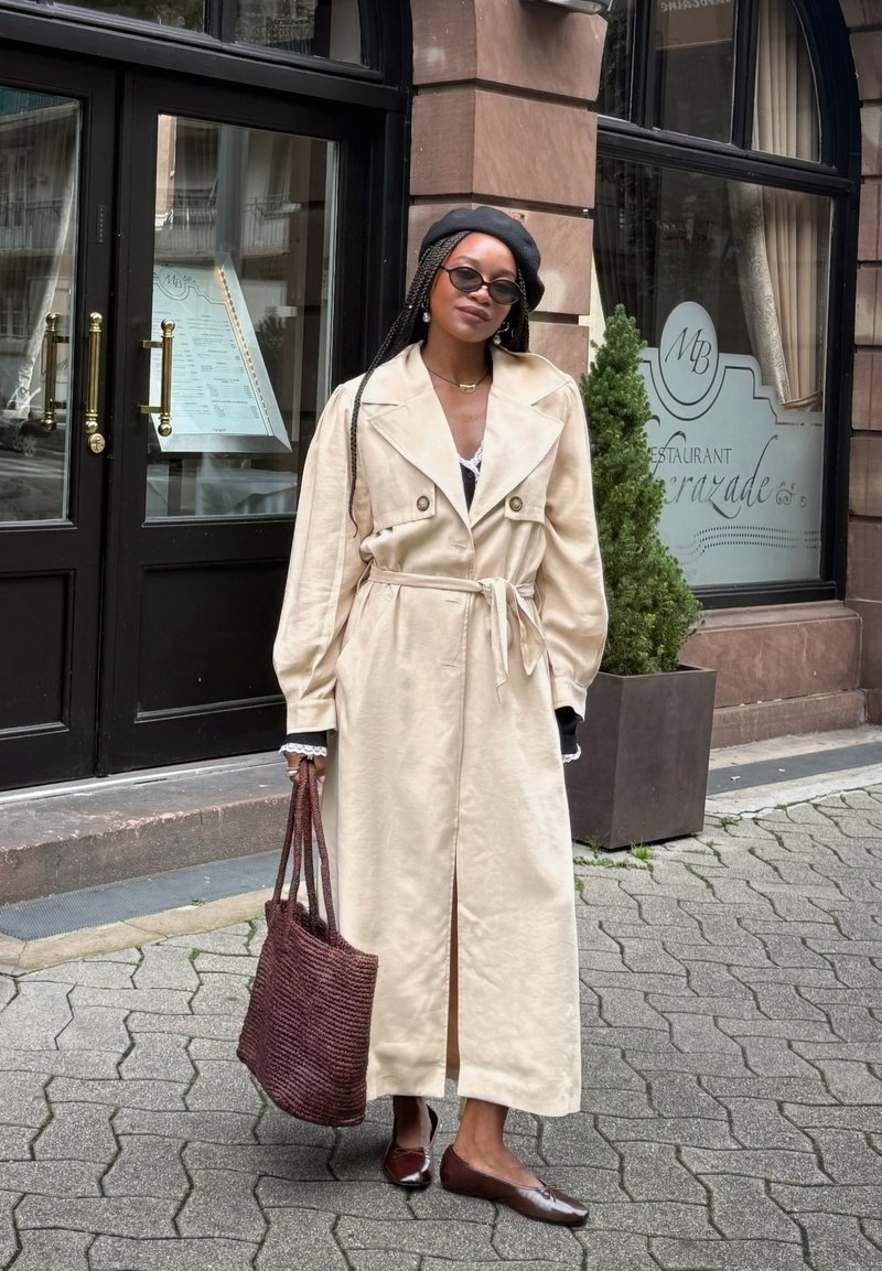 Casaco trench bege com cinto, lapelas oversized e detalhes de bolso. Bolsa tote bordô tecida e sapatilhas castanhas. Fundo de pavimento em tijolo e pedra.