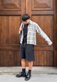 Jeune homme debout devant une grande porte en bois portant une chemise à carreaux, un short noir, des chaussures noires et un sac à bandoulière.
