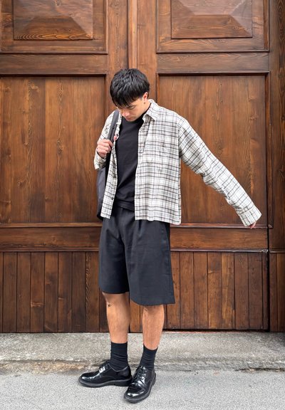 Joven de pie frente a una gran puerta de madera, vestido con una camisa de cuadros, pantalones cortos negros, zapatos negros y llevando una bolsa al hombro.